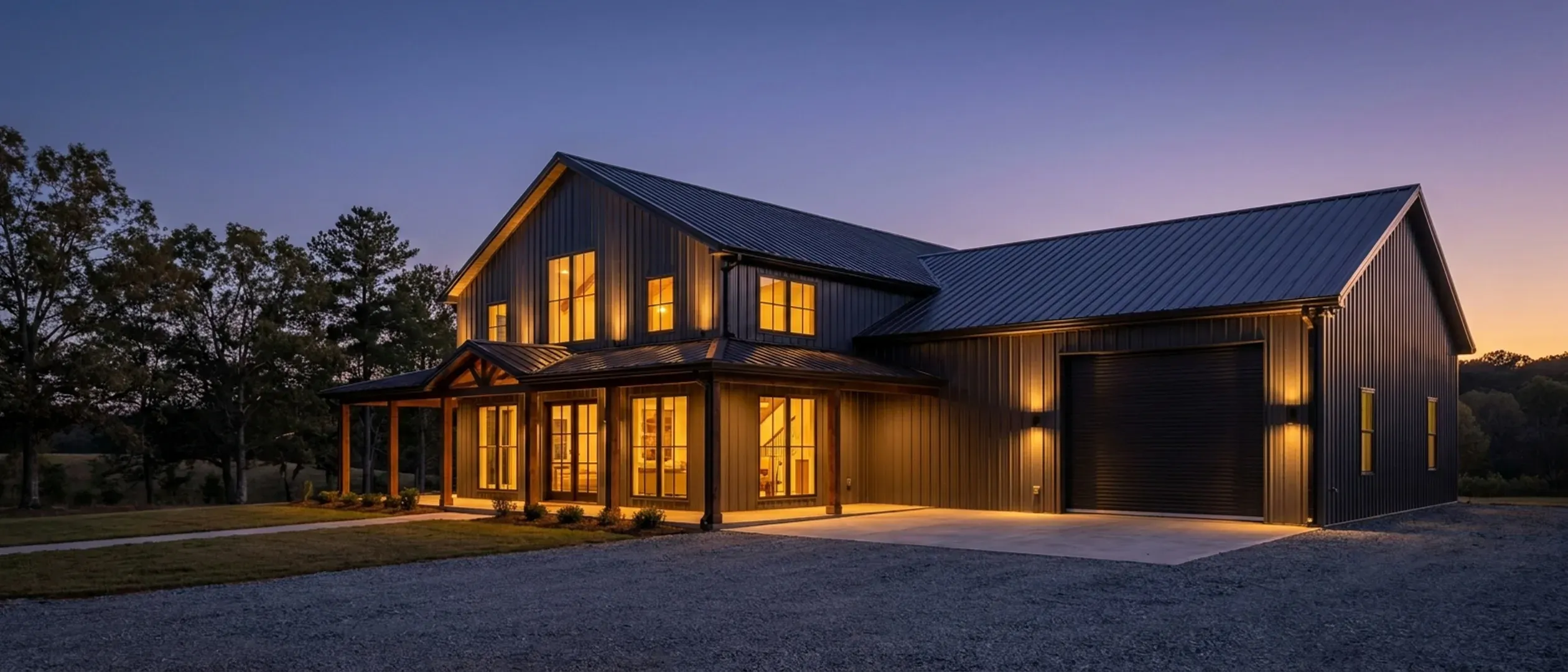 A barndominium at dusk with warm light glowing from every window and exterior lighting along the porch and roofline against a deep blue twilight sky.